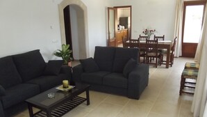 A living room with a dark-colored sofa set, a coffee table, and a potted plant. There is a dining area with wooden chairs and a table visible through an open doorway. - EXCLUSIVE VILLA WITH PRIVATE POOL, JUST 1 MINUTE FROM CALÓ DES POU BEACH (Cala D'or/Santanyi)
