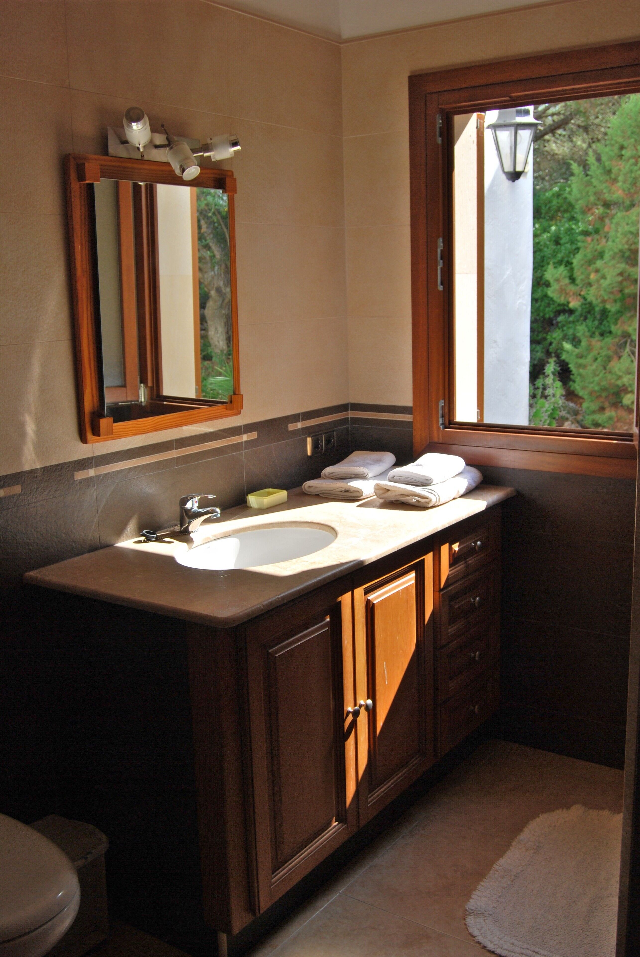 A bathroom with a wooden vanity, two sinks, a mirror, and a window with a view of greenery.