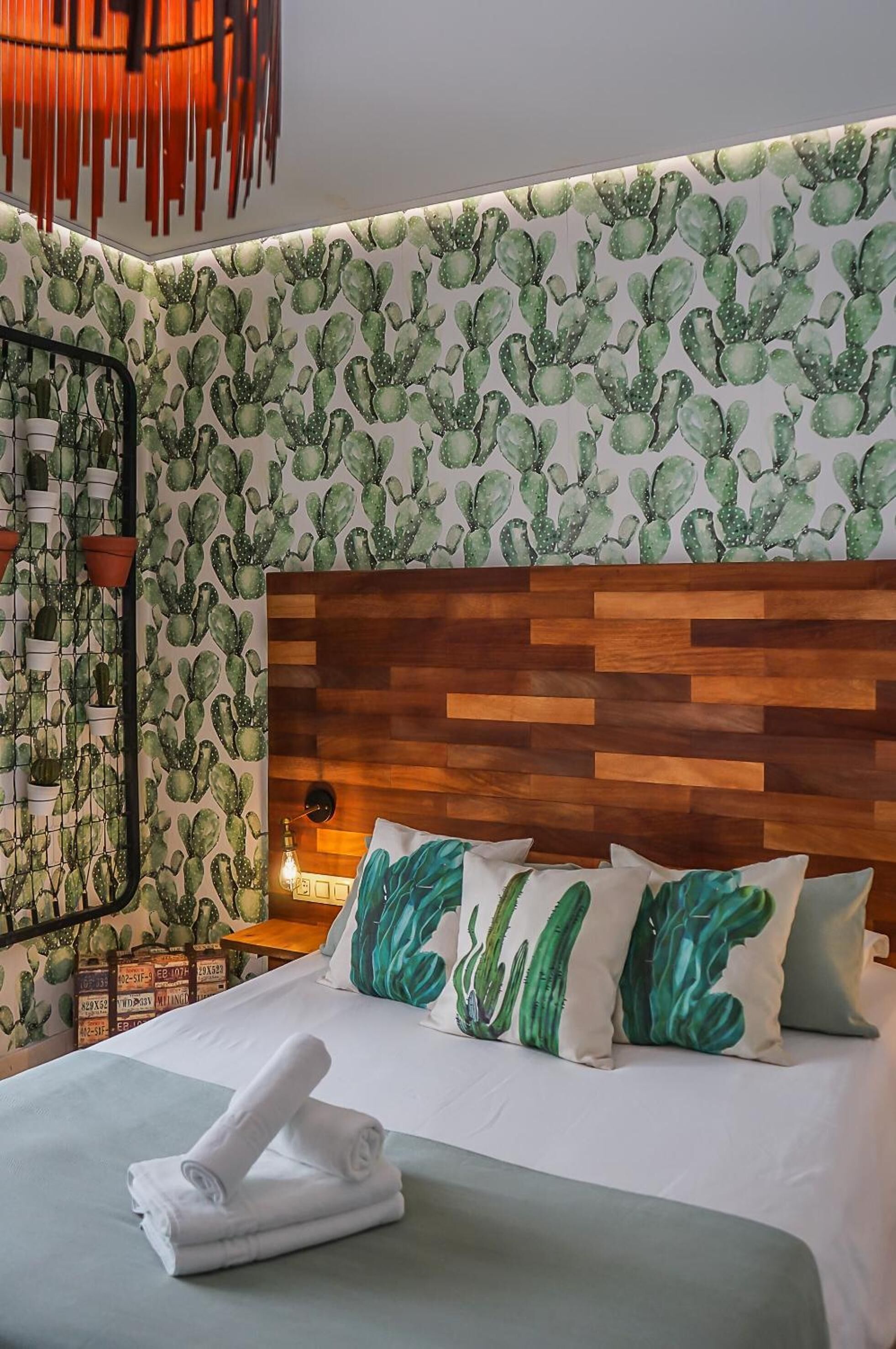 A hotel room with a cactus-themed wallpaper, a wooden headboard, and a neatly made bed with green and white pillows.