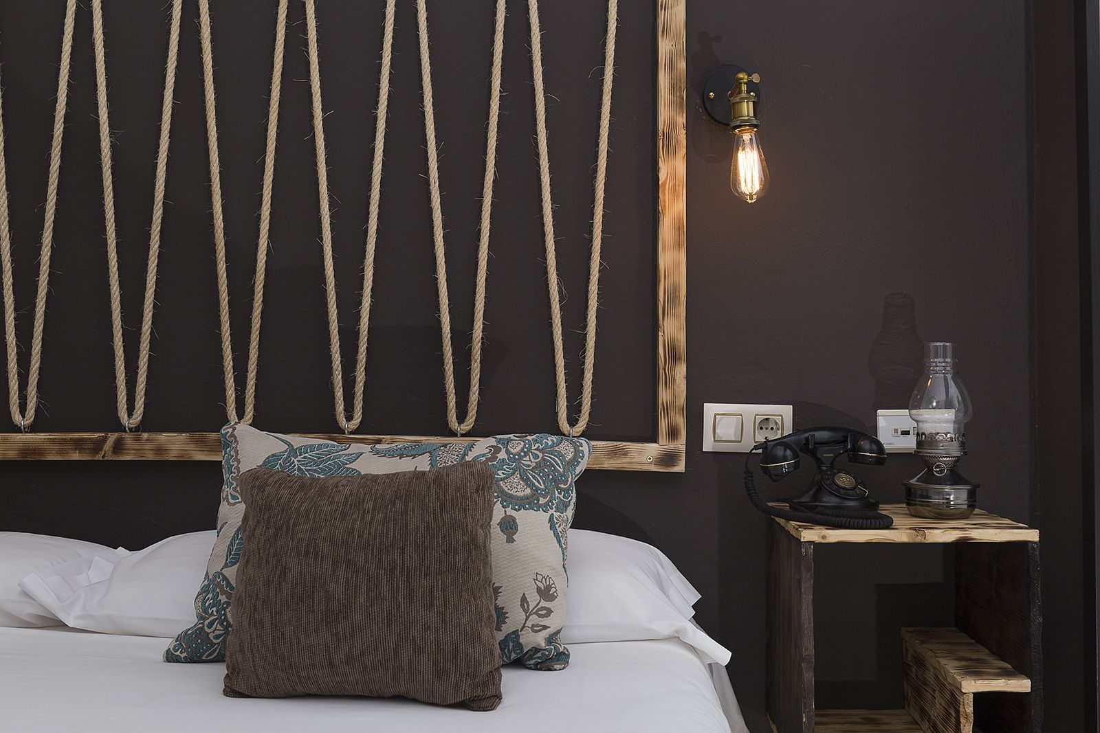 A bedroom with a bed, a headboard with ropes, a nightstand with a lamp, and a wall socket.