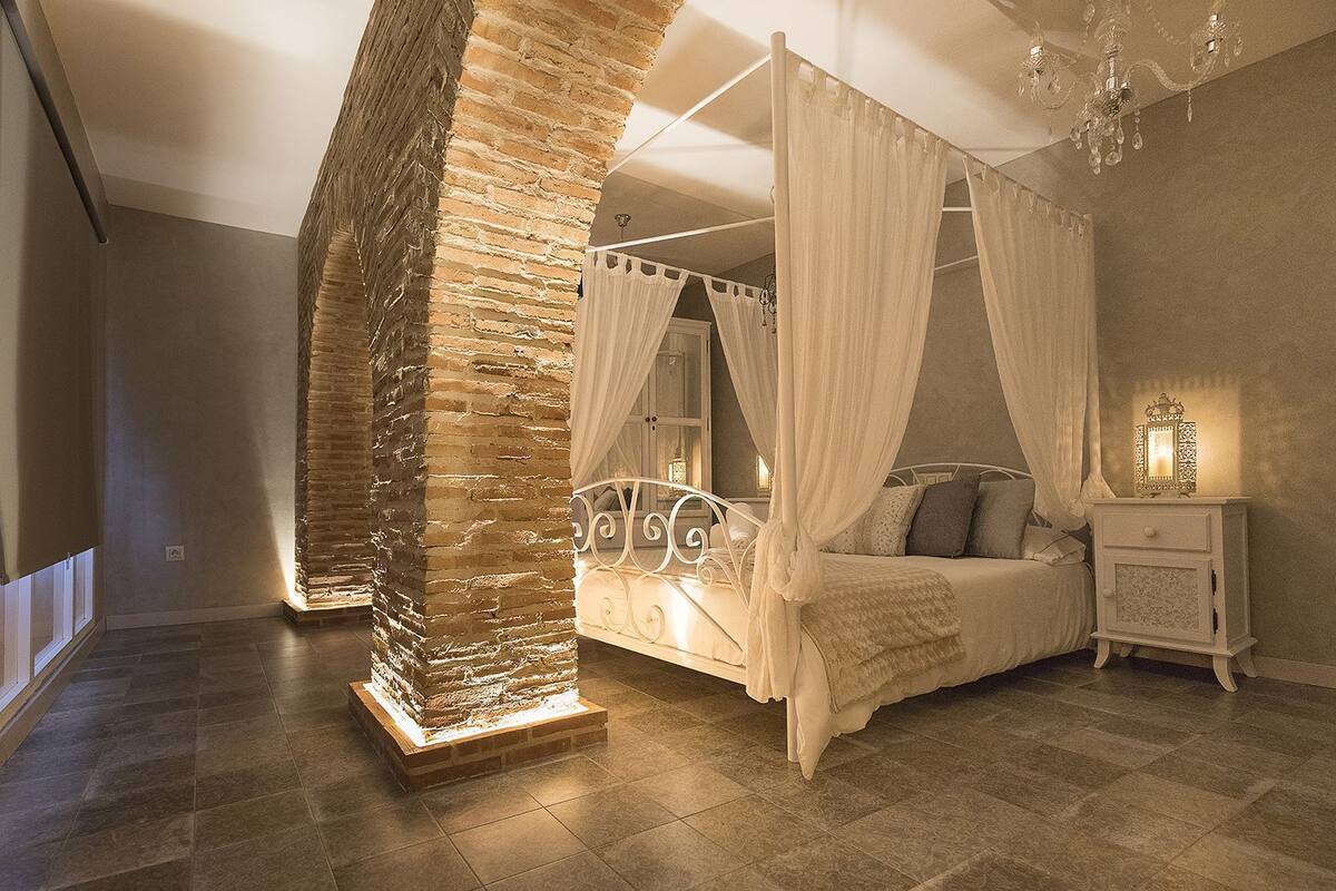 Een slaapkamer met een hemelbed, nachtkastje, decoratieve zuil en een kroonluchter.