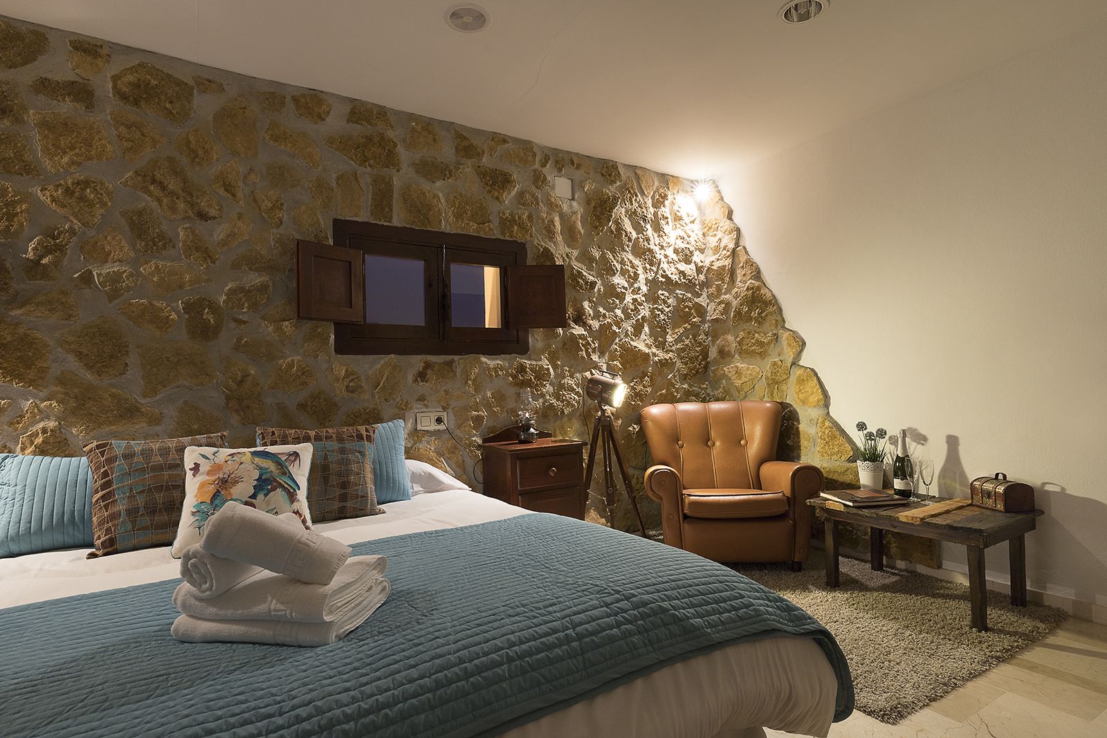 A bedroom with a stone wall, a bed with blue bedding, a leather armchair, a small table with a lamp, and a nightstand.