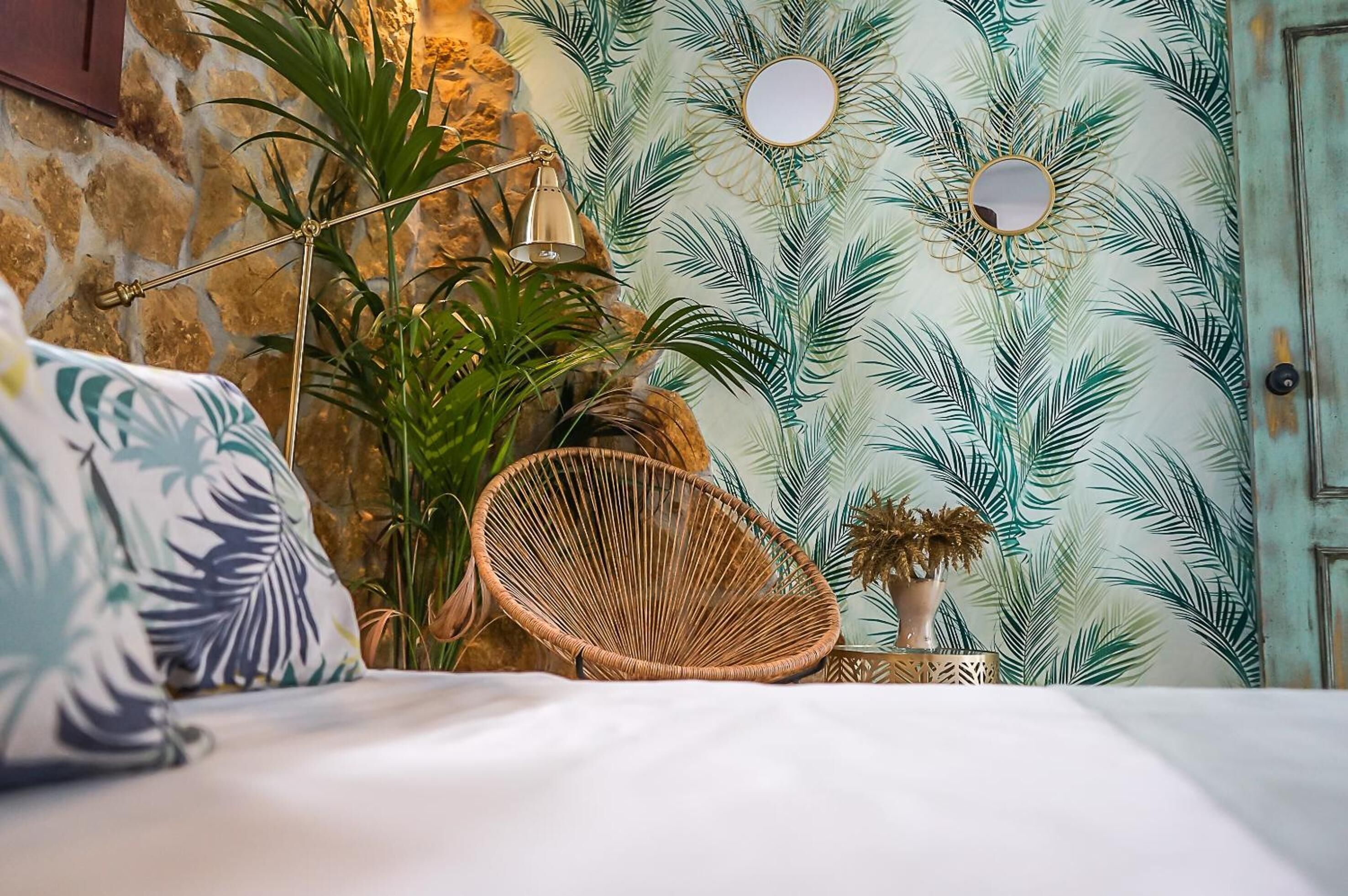 Une chambre avec un mur en pierre, un lit avec une literie blanche, une chaise en osier et un mur orné d’un motif de feuilles de palmier.