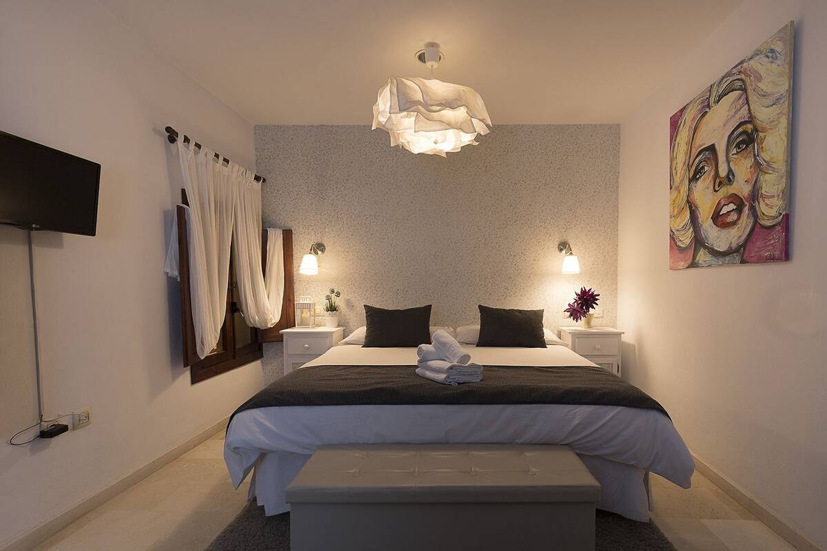Een slaapkamer met een bed, nachtkastjes, een televisie, een schilderij en een kroonluchter.