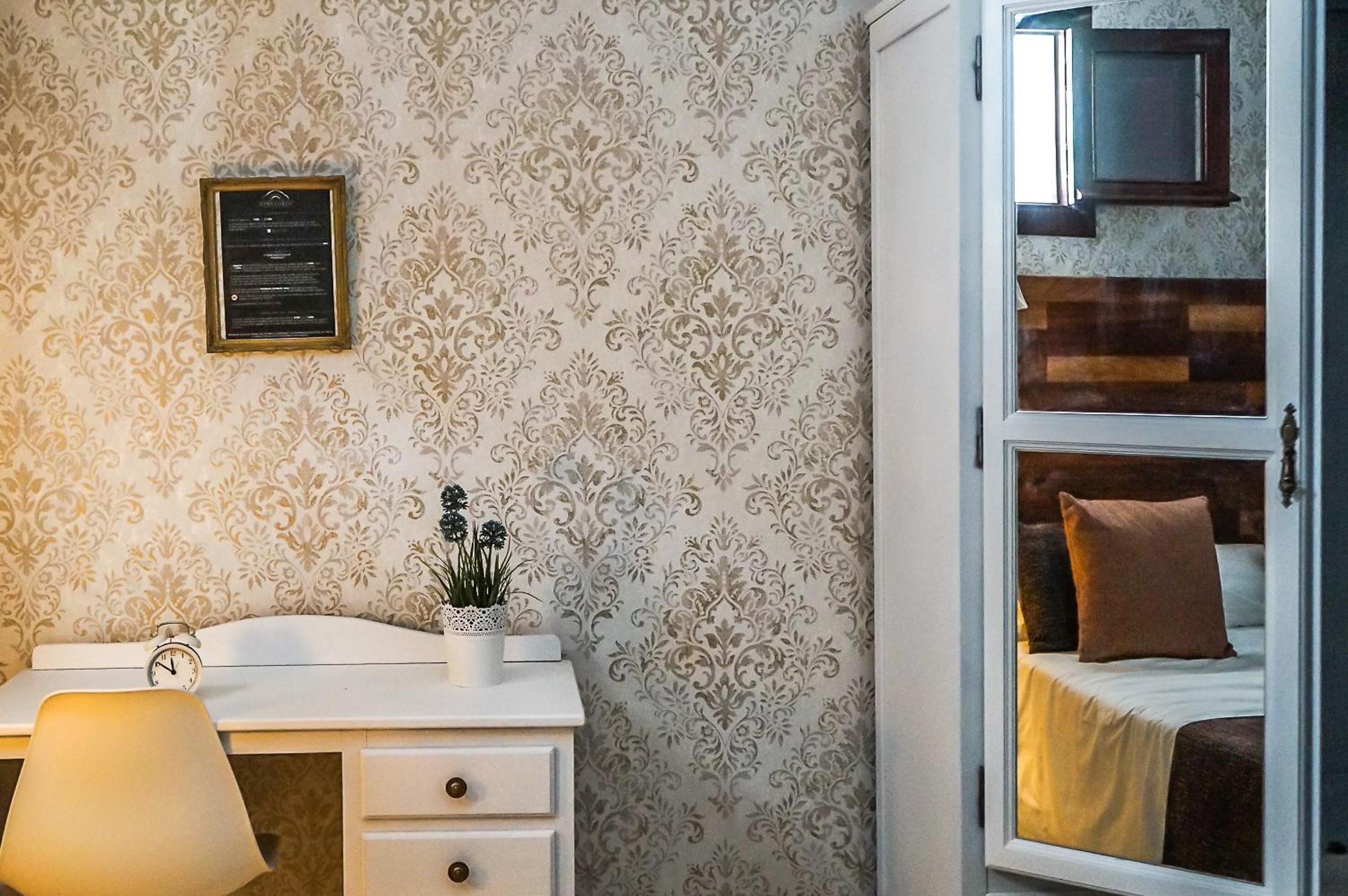 Un dormitorio con cama, una mesita de noche con lámpara, un diploma enmarcado en la pared y una ventana que da a otra habitación.