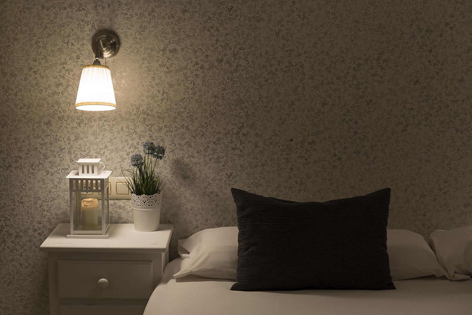 A bedroom with a nightstand, a wall-mounted lamp, a small table with a candle and a plant, and a bed with a dark pillow.
