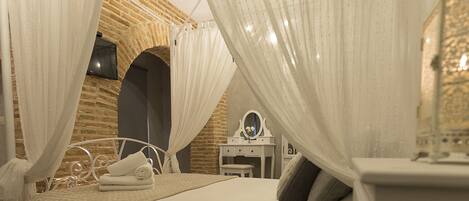 Un dormitorio con pared de ladrillo, cama con ropa blanca, mesita de noche con espejo y cama con dosel y cortinas traslúcidas.
