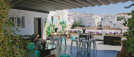 An outdoor seating area with tables and chairs, a decorative wall with tropical leaves, and a view of white buildings in the background.