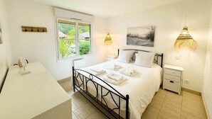 A bedroom with a bed, bedside tables, a desk, and a window with a view of greenery. - Villas La Clairière aux Chevreuils (Moliets-et-Maa)