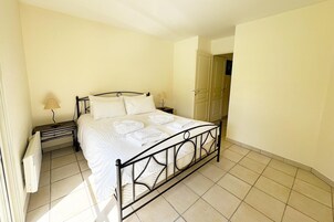 Un lit bien fait, avec du linge de lit blanc et des oreillers, une table de chevet avec une lampe, et une porte qui donne sur une autre pièce.