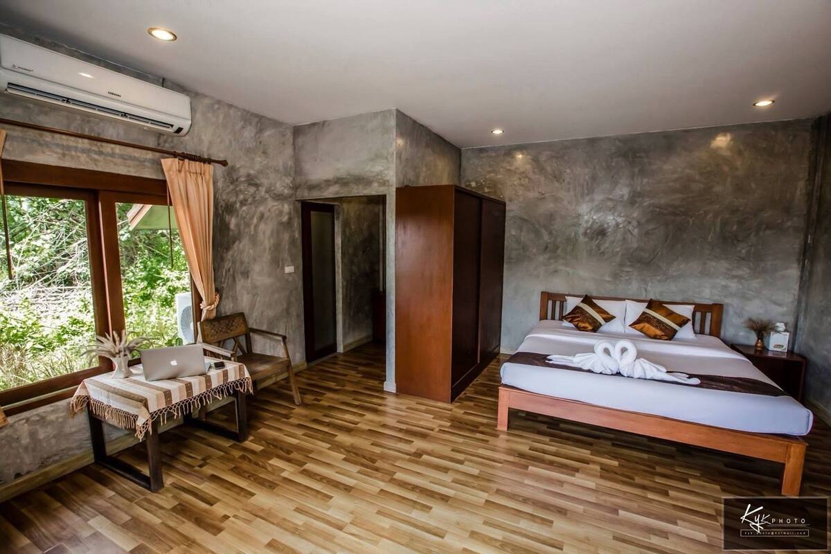 A bedroom with a wooden floor, a bed with a white bedspread and decorative pillows, a bedside table with a laptop, and a window with curtains.