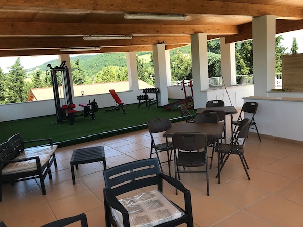 A covered outdoor area with exercise equipment, a dining table with chairs, and a view of a green field and trees. - Colle Sul Lago (Cingoli)