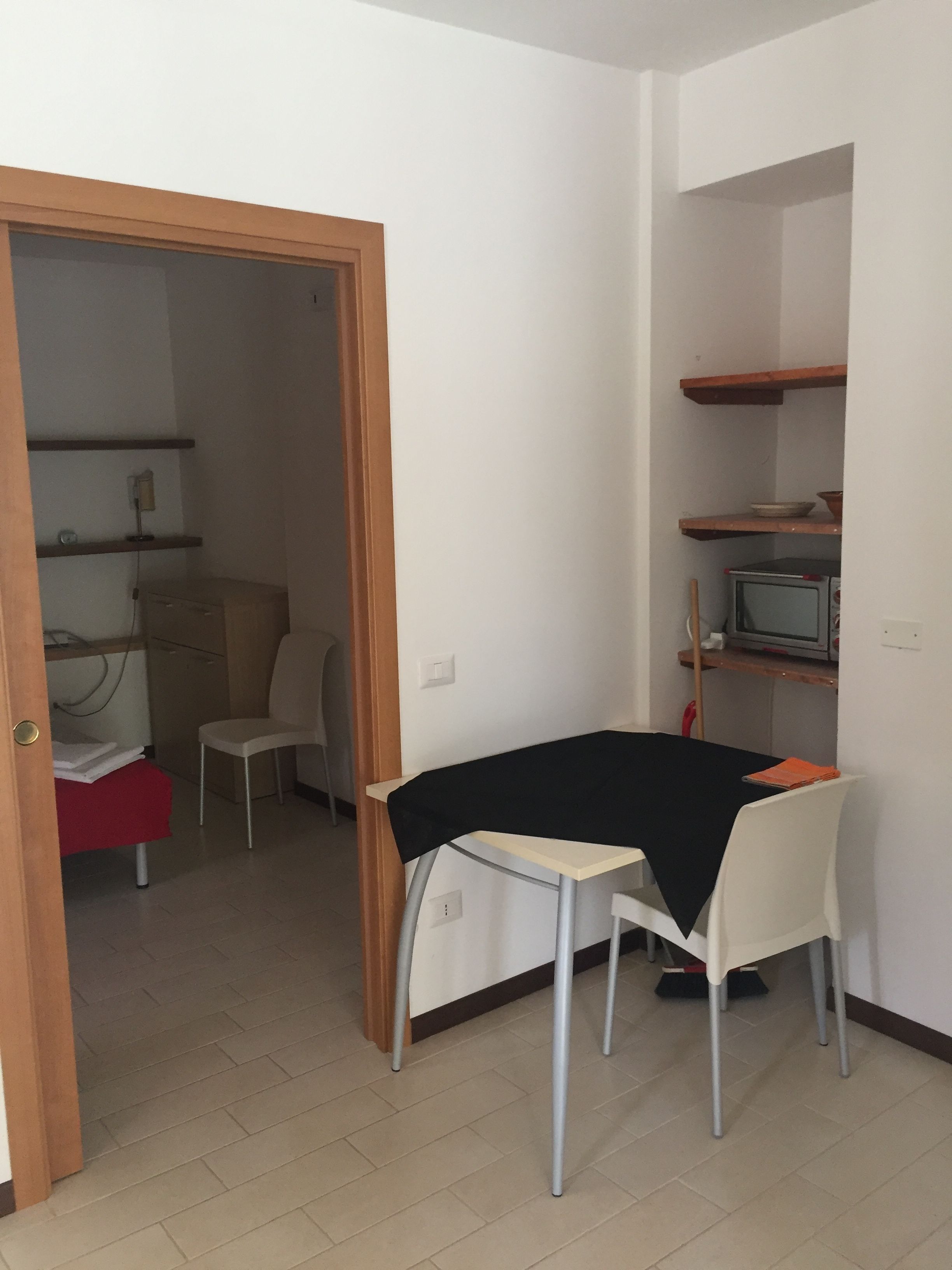 A small, well-lit room with a dining table and chairs, a television on a shelf, and a door leading to another room.