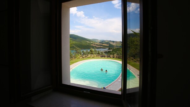 A view through a window of a swimming pool with two people in the water, surrounded by greenery and hills. - Colle Sul Lago (Cingoli)