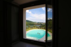A view through a window of a swimming pool with two people in the water, surrounded by greenery and hills. - Colle Sul Lago (Cingoli)