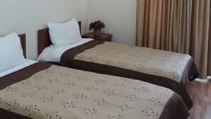 A hotel room with two beds, a nightstand with a vase of flowers, an air conditioning unit, and a window with curtains. - Hotel Rio (Tbilisi)