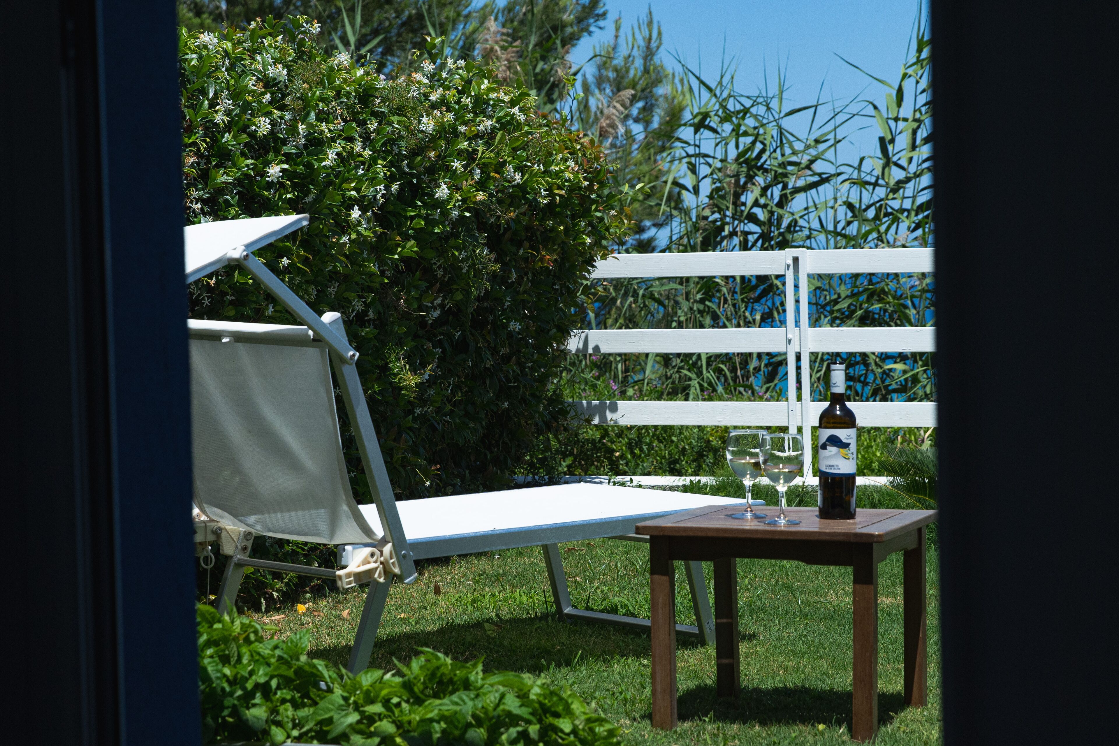 A garden patio with a white chair, a small table with a bottle of wine and two glasses, and a white fence in the background.