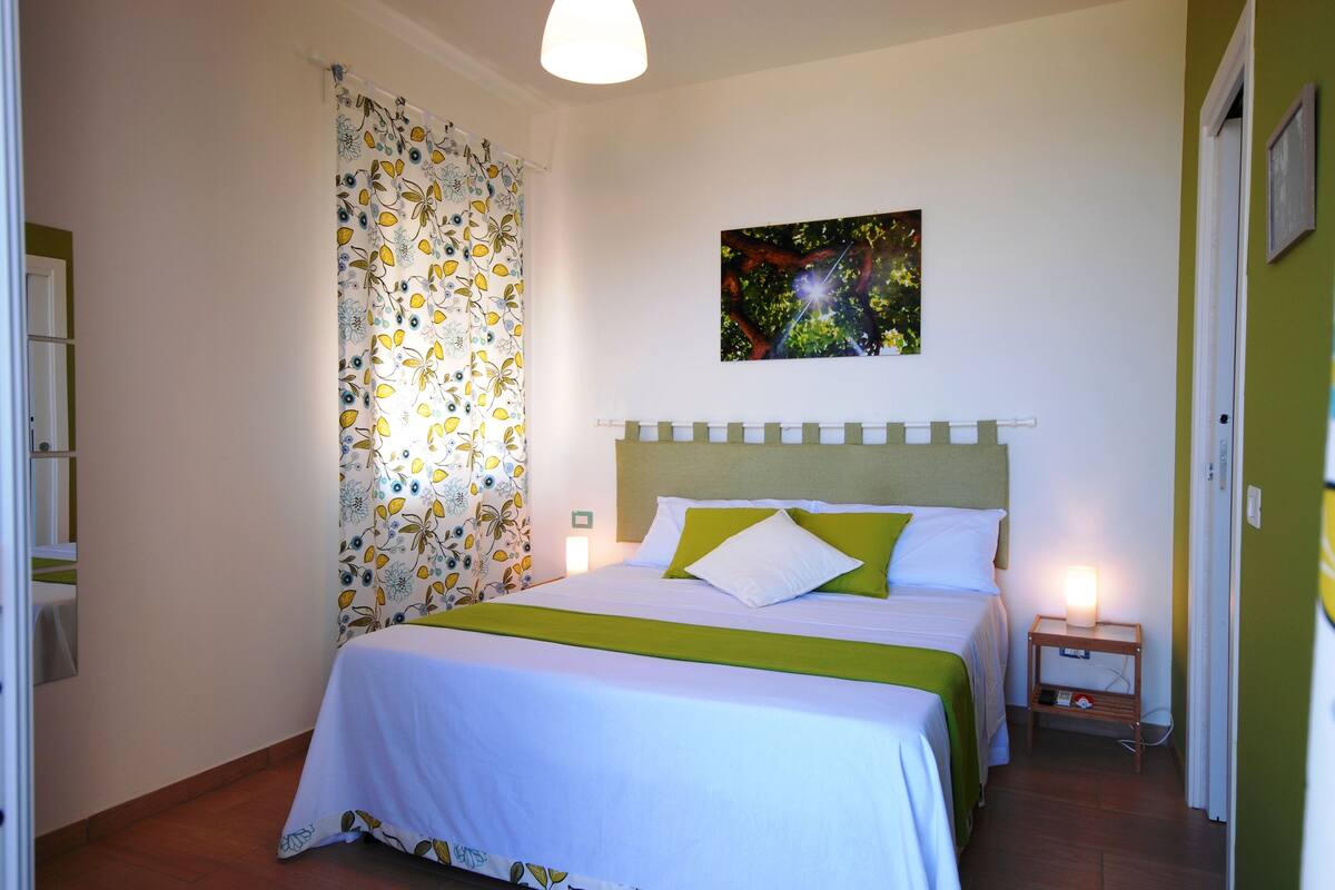 A neatly arranged bedroom with a bed, a bedside table, a nightstand, a wall-mounted picture, and a curtain with a floral pattern.