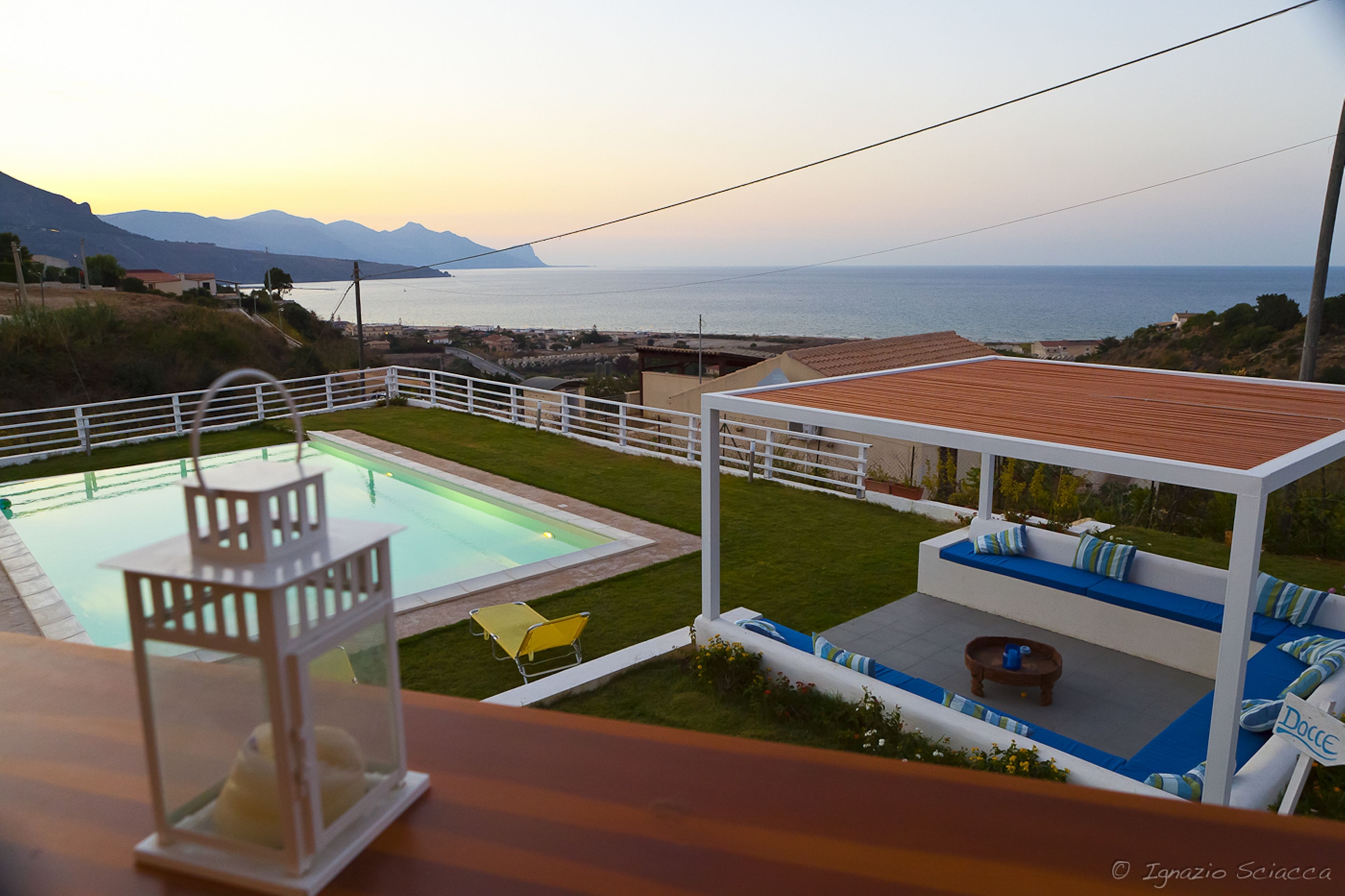 Un espace piscine agrémenté d’une clôture blanche, d’une petite terrasse avec des sièges, et offrant une vue sur la mer et les montagnes.