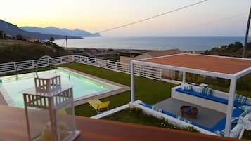 Un espace piscine agrémenté d’une clôture blanche, d’une petite terrasse avec des sièges, et offrant une vue sur la mer et les montagnes.