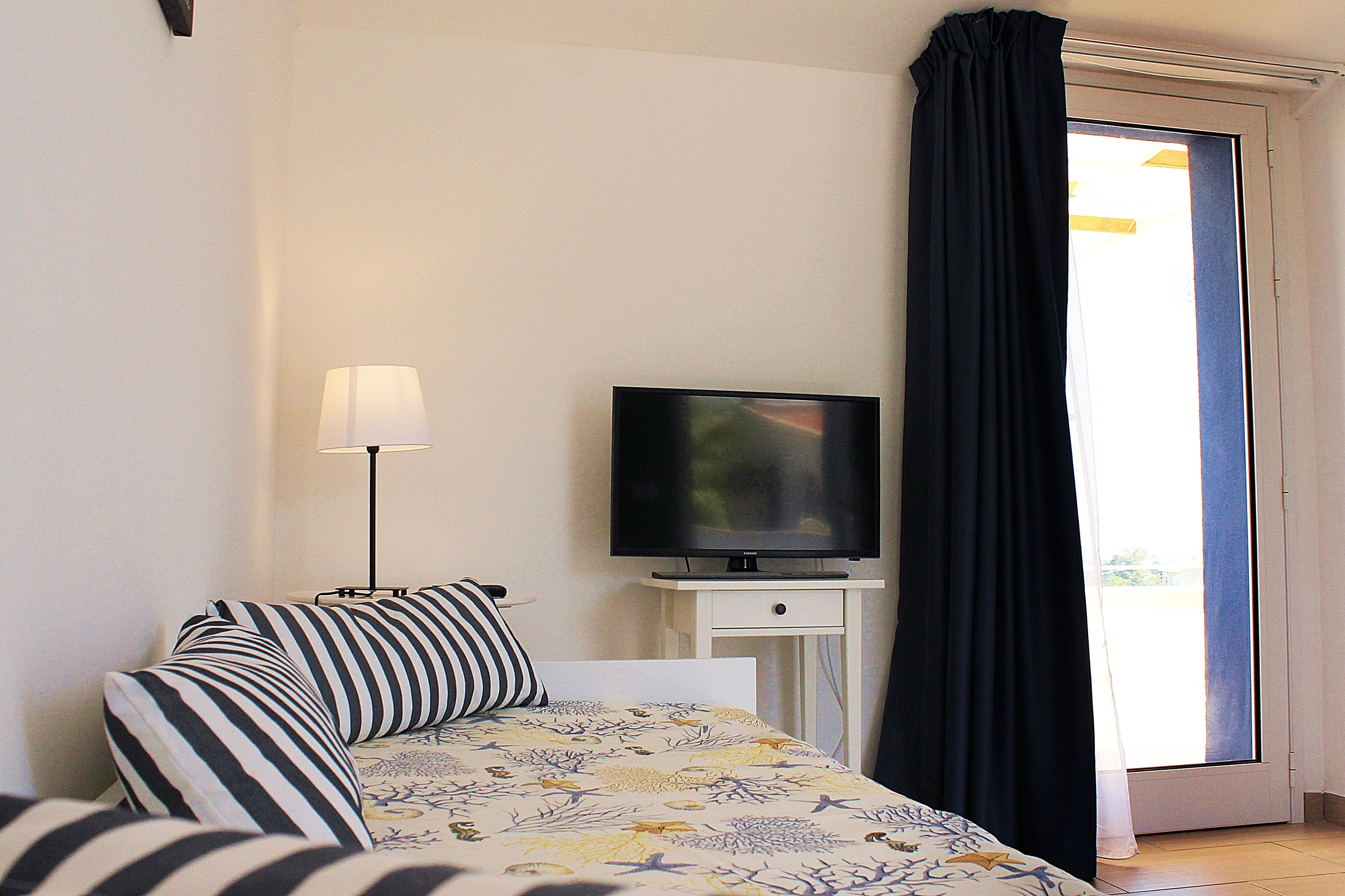 Un dormitorio con cama, televisor sobre un mueble, lámpara y vistas al exterior a través de una ventana con cortinas.