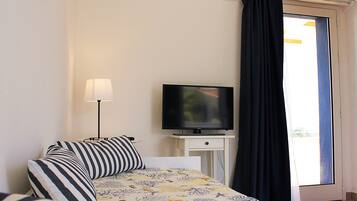 Un dormitorio con cama, televisor sobre un mueble, lámpara y vistas al exterior a través de una ventana con cortinas.