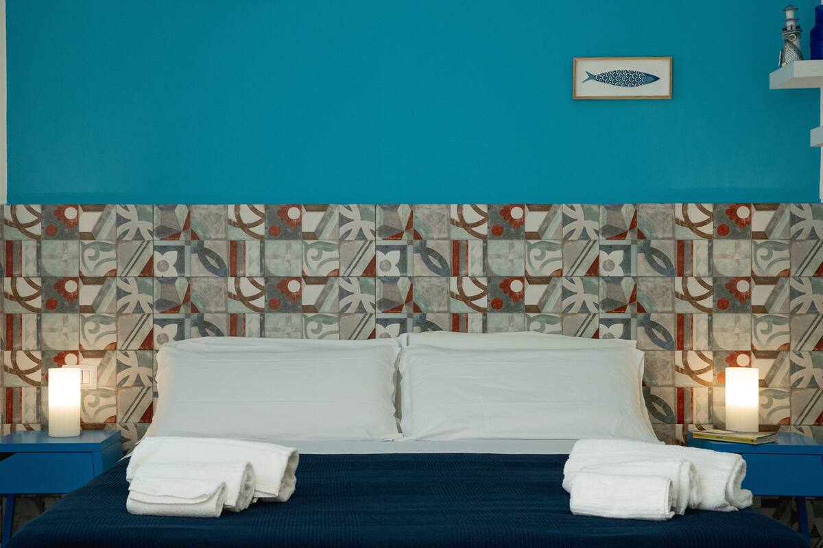 A hotel room with a patterned headboard, a bed with white linens, two bedside tables with lamps, and a framed picture on the wall.