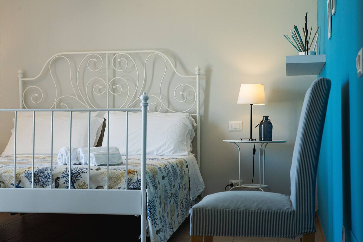 A neatly made bed with a white metal headboard, a bedside table with a lamp, and a chair.