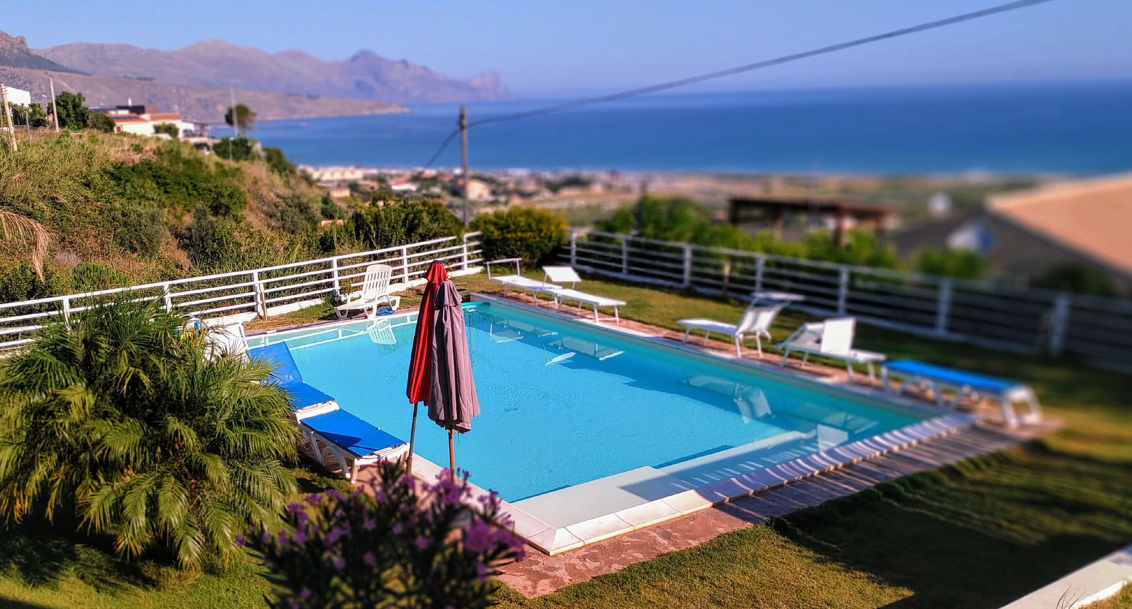 A swimming pool with lounge chairs and umbrellas, surrounded by greenery and a view of the sea and mountains.