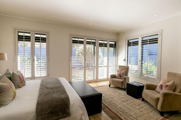A bedroom with a large bed, two armchairs, a small table, and large windows with blinds.