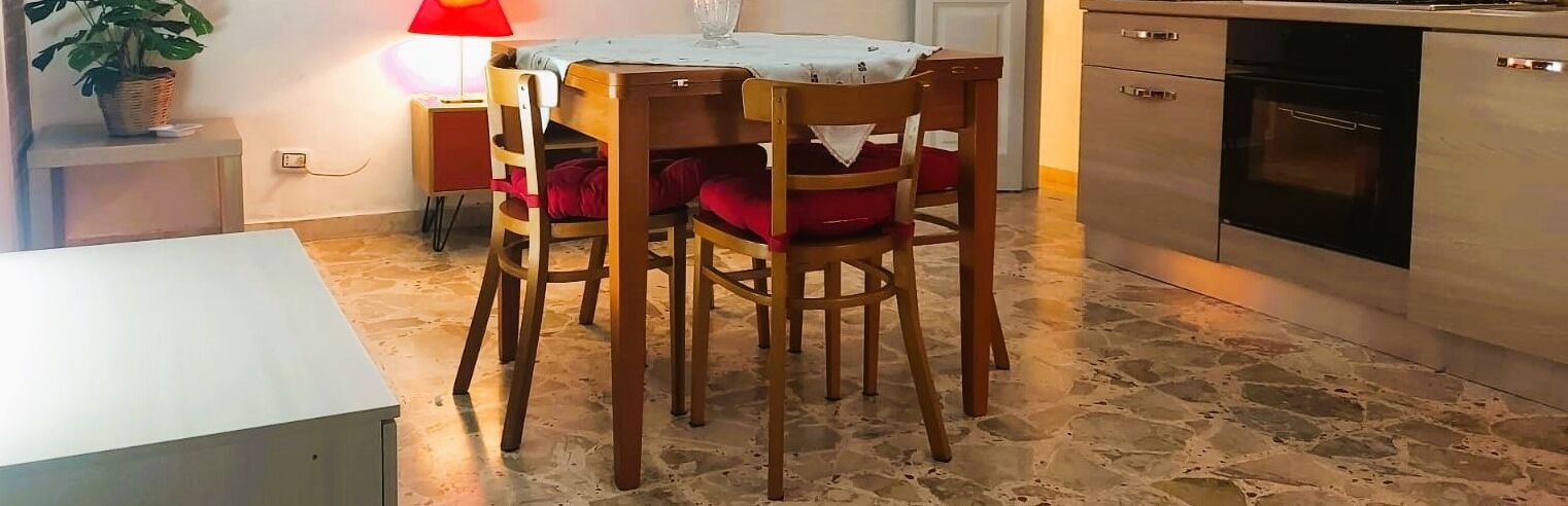 A dining area with a wooden table and chairs, a red cushion, a lamp, and a kitchen with a stove and oven.