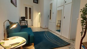 Una sala de estar con un sofá azul, una mesa de centro con papeles, una lámpara, una alfombra y una estantería.
