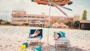 A beach with two lounge chairs under a striped umbrella, a building with a sign in the background, and a clear sky.