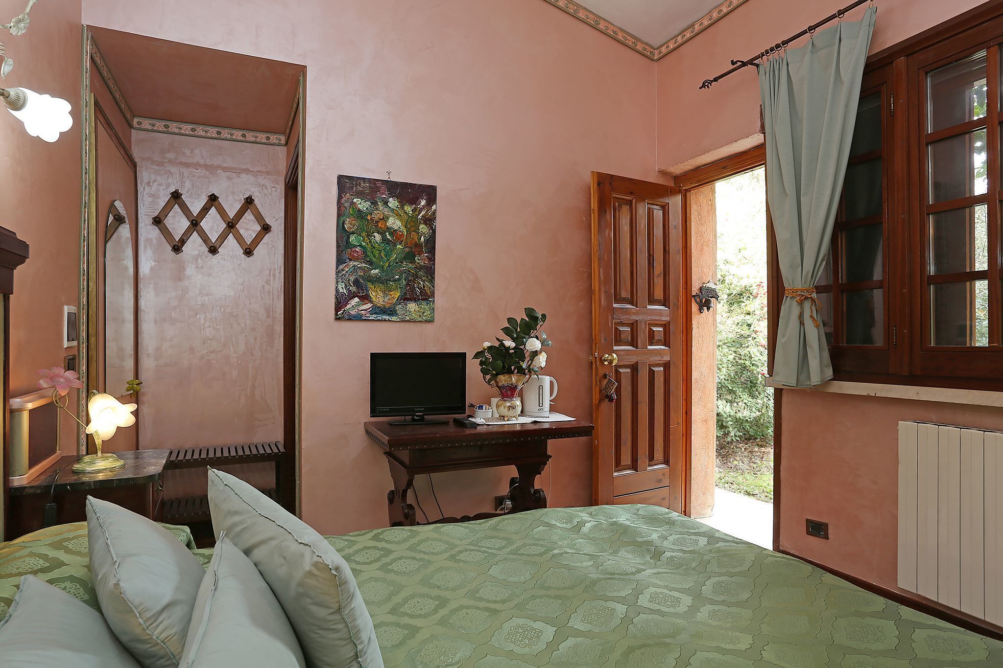 A bedroom with a bed, a desk with a computer, a mirror, a painting, and a door leading outside.