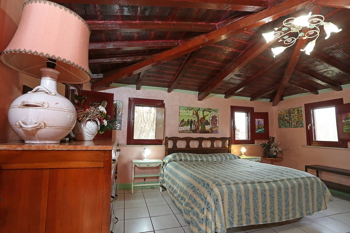 Un dormitorio con techo de madera, una cama con cubrecama estampado, una mesita de noche con lámpara y un jarrón grande sobre un aparador.