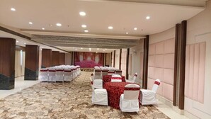 Uma sala de banquetes com mesas e cadeiras dispostas em fileiras, piso coberto de carpete e palco com um painel floral.