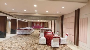 A banquet hall with rows of tables and chairs, a carpeted floor, and a stage with a floral backdrop. - Hotel Grand Meridian (Katni)
