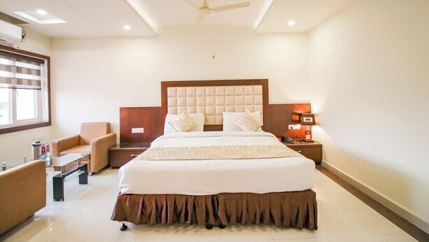 A hotel room with a large bed, a desk, a chair, a nightstand, and a ceiling fan. - Hotel Grand Meridian (Katni)