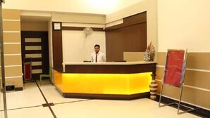 A reception desk with a person standing behind it, a clock on the wall, and a red sign on the right. - Hotel Grand Meridian (Katni)