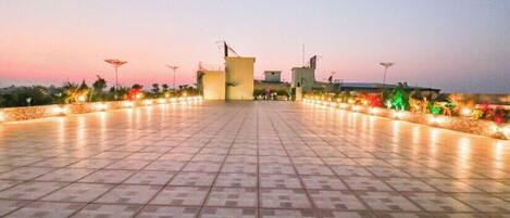 A spacious outdoor area with a tiled floor, a few buildings in the background, and decorative lights along the edges.