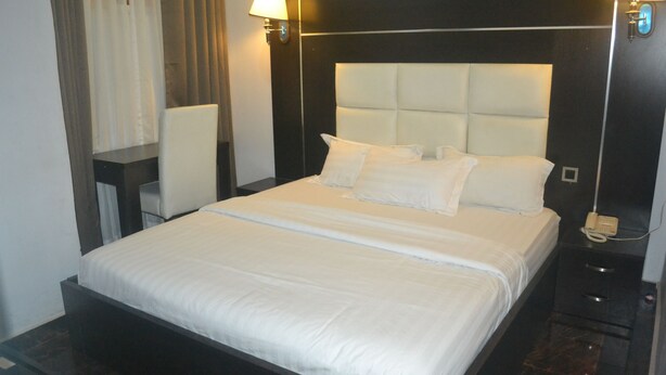 A hotel room with a large bed, a bedside table, a chair, and a nightstand with a phone. - Mac Dove Lounge & Suites ltd (Lagos)