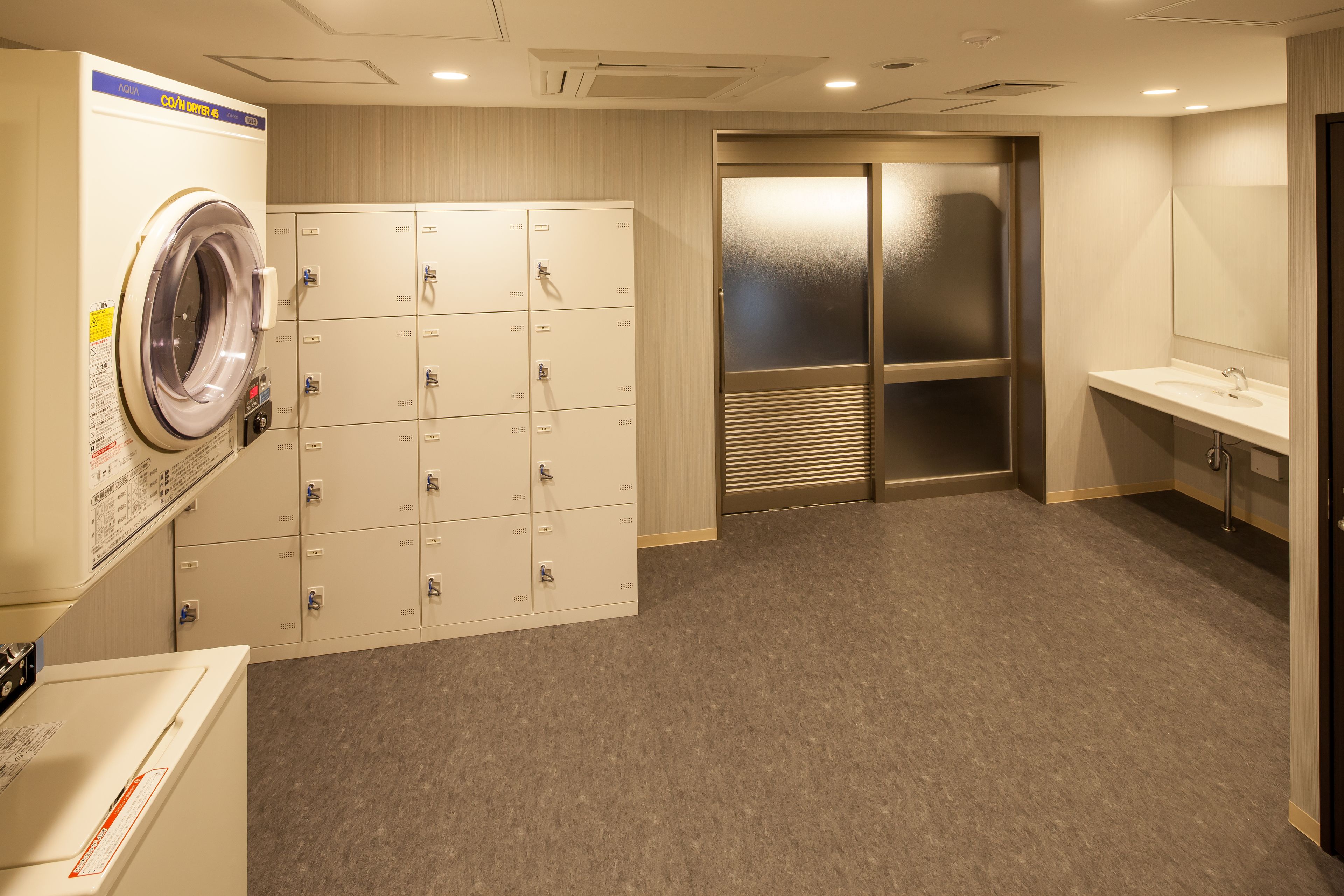 A hotel room with a washing machine, a row of lockers, a sink, and a mirror.