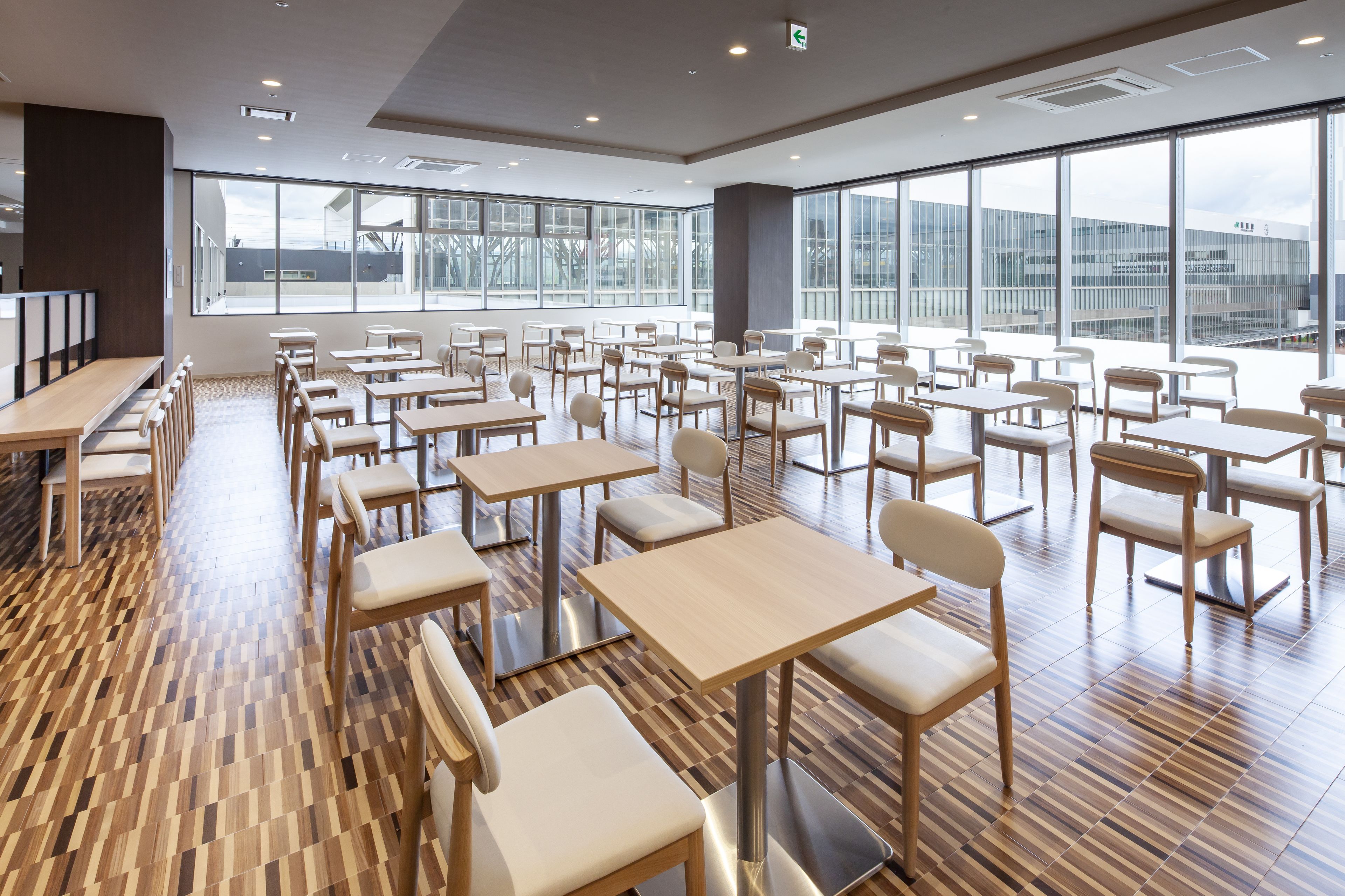 A spacious dining area with multiple tables and chairs, large windows offering a view of the outside, and a polished wooden floor.