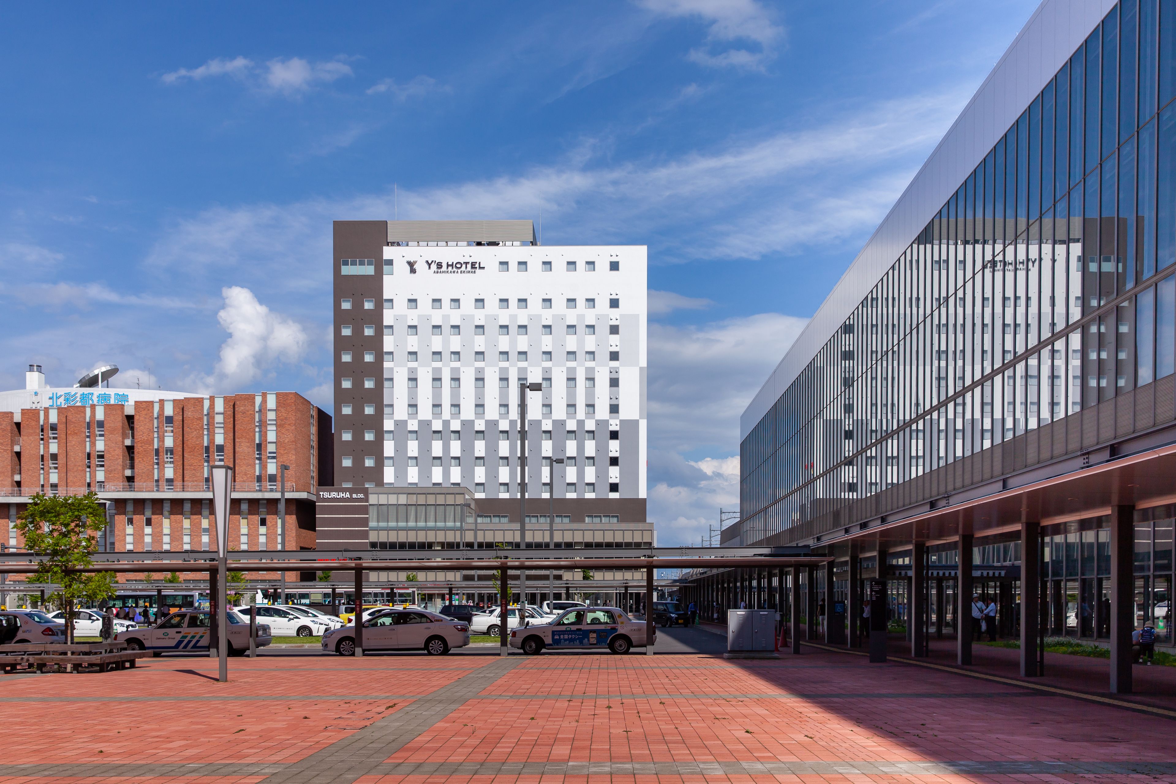 A modern hotel with a glass facade, a parking area with several cars, and a clear blue sky.