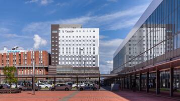 A modern hotel with a glass facade, a parking area with several cars, and a clear blue sky.
