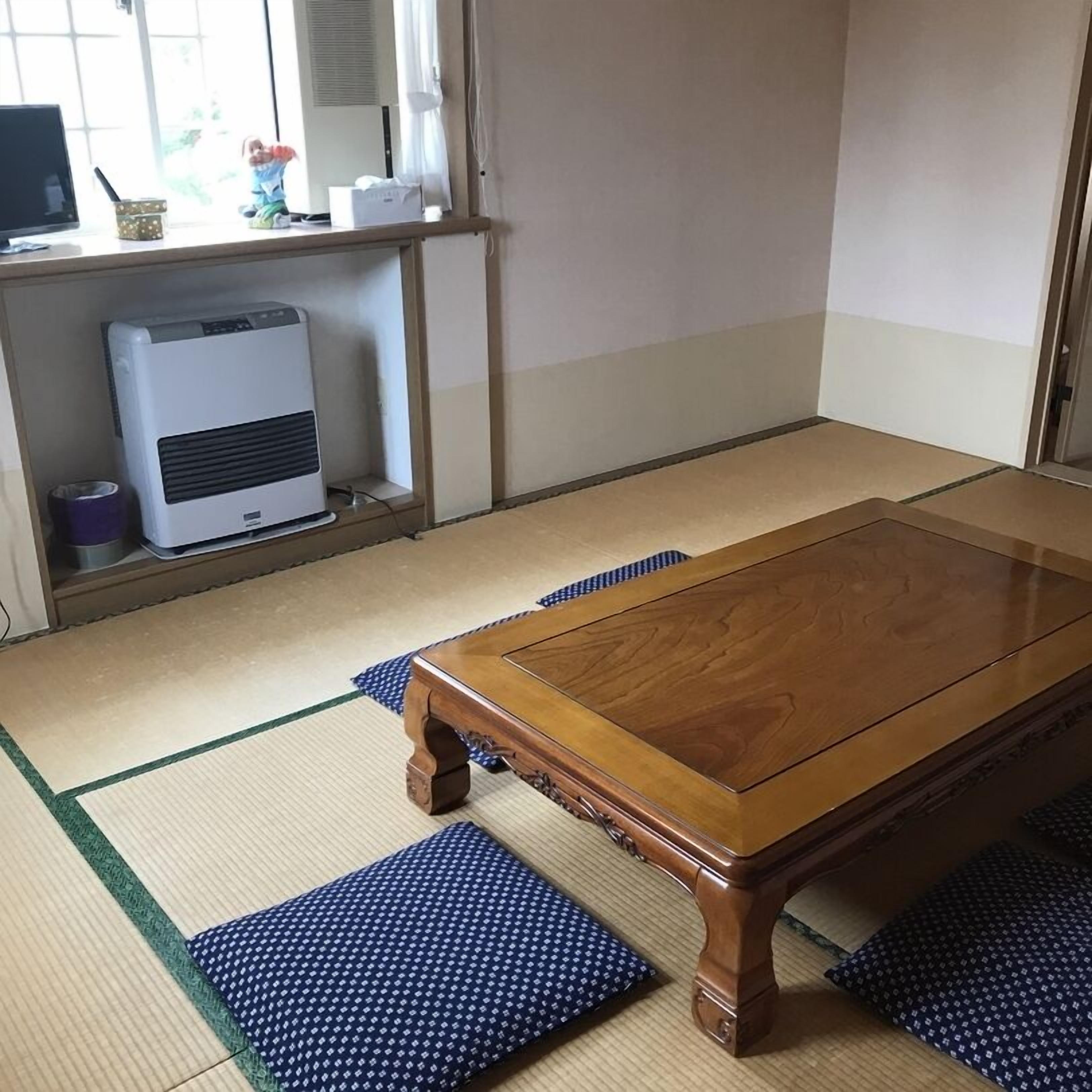 A traditional Japanese tatami room with a wooden table, tatami mats, a heater, and a TV on a shelf.