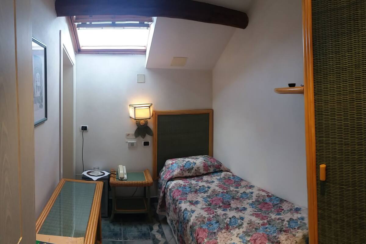 A small, narrow room with a bed, a desk, a wall-mounted lamp, and a window.