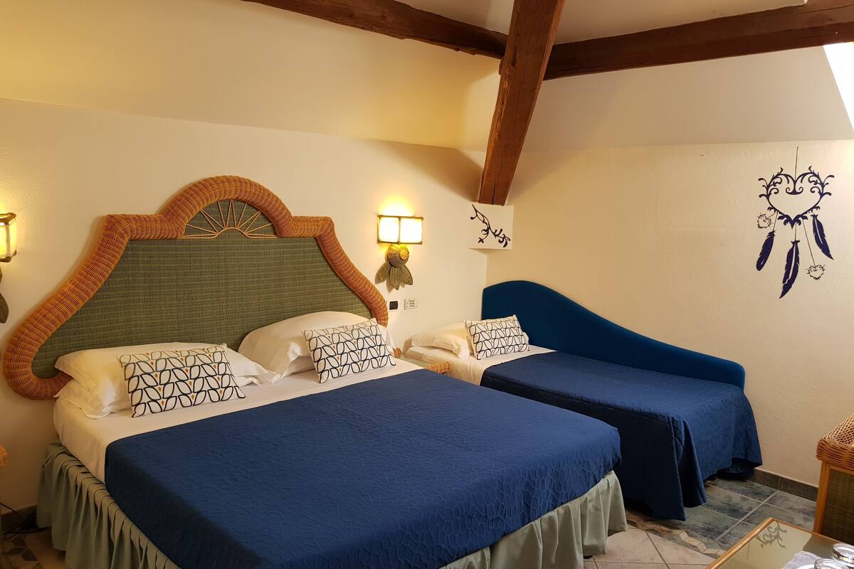 A hotel room with two beds, each with a blue bedspread and matching pillows. There is a wall-mounted lamp and a dreamcatcher decoration.