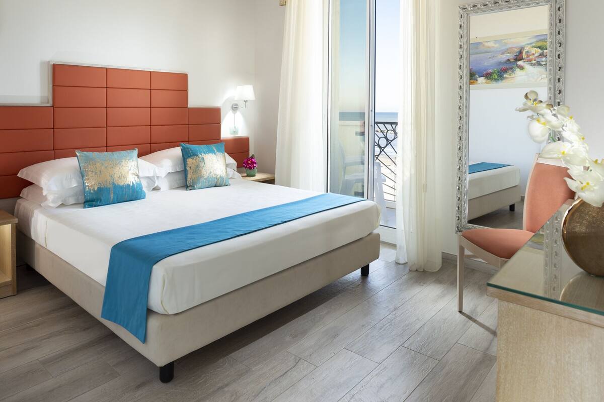 A hotel room with a large bed, a nightstand, a lamp, a chair, a mirror, and a balcony with a railing.