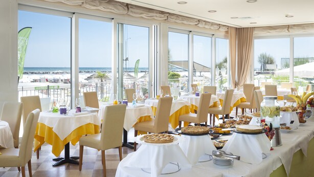 Un restaurante con mesas dispuestas para comer, un bufé con diversas opciones de comida y vistas a la playa y al océano a través de amplias ventanas.